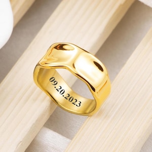 May include: A polished gold-tone ring with a wide, irregular, wavy band. The interior of the ring is engraved with the date 09.20.2023 in a clear, dark font. The surface of the metal is smooth and reflective, catching light across its curved edges.