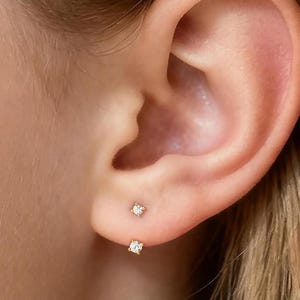 Könnte beinhalten: Nahaufnahme eines Ohrs mit zwei kleinen, goldfarbenen Ohrsteckern. Der obere Ohrring ist ein kleiner, runder, klarer Stein. Der untere Ohrring ist ein quadratischer, klarer Stein.