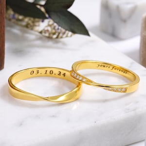 May include: Two gold-toned rings with a twisted band design. One ring features the engraved date 03.10.24 on the inner surface. The other ring displays the engraved text yours forever and is accented with a row of small, clear, sparkling stones.