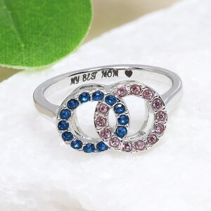 May include: A silver ring with two interlocking circles, one with blue and the other with pink gemstones. The band is engraved with "MY BEST MOM" and a small heart. The ring is displayed on a white, textured surface.