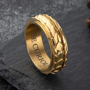 May include: A gold-colored ring with a textured, geometric pattern. The ring has the words "BE CURIOUS" engraved on the inside. The ring is displayed on a dark, textured surface.