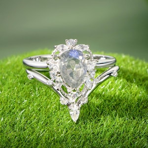 May include: A silver engagement ring set with a pear-shaped, iridescent gemstone, surrounded by smaller, clear stones. The ring sits atop a bed of green moss, showcasing its intricate design and sparkle.