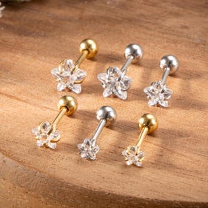 May include: Six star-shaped stud earrings. Three are gold-colored with clear crystal star accents, and three are silver-colored with clear crystal star accents. The earrings are displayed on a wooden surface.