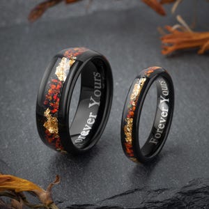 May include: Two black rings with a band of red and gold flecks. The rings have the words "Forever Yours" engraved on the side. The rings are displayed on a dark gray surface.