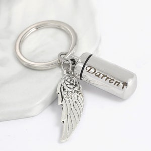 May include: Silver keychain with a small cylindrical urn engraved with the name "Darren". The urn is attached to a silver angel wing charm with a rose detail.