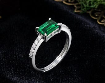 Puede incluir: Un anillo de tono plateado con una piedra preciosa verde de corte esmeralda rectangular en el centro, flanqueada por una hilera de pequeñas piedras transparentes de corte redondo a lo largo de la banda. El anillo tiene un acabado pulido y una abertura.