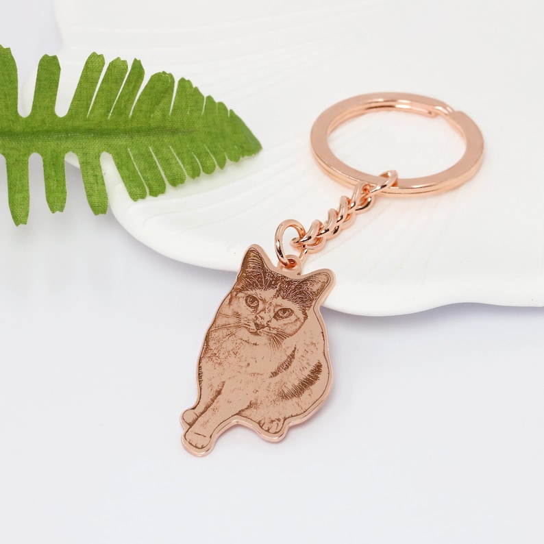 May include: Rose gold keychain with a personalized engraved image of a cat. The cat is sitting with its head tilted and looking at the camera.