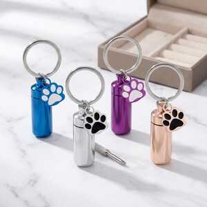 Op de afbeelding: Vier cilindervormige metalen urn-sleutelhangers in blauw, zilver, paars en ros&eacute;goud. Elke sleutelhanger heeft een ring en een bedel in de vorm van een pootafdruk. Een kleine metalen schroevendraaier ligt bij de zilveren sleutelhanger.