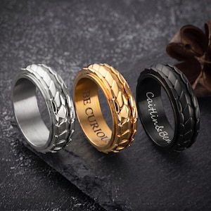 Puede incluir: Tres anillos giratorios texturizados en plata, oro y negro. El anillo dorado tiene grabado "BE CURIOUS" en el interior. El anillo negro está grabado con "Caitlin & Ben". Los anillos se muestran sobre una superficie oscura.