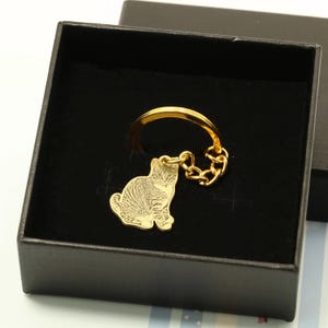 May include: Gold-colored keychain with a small, engraved image of a cat. The keychain is in a black gift box.