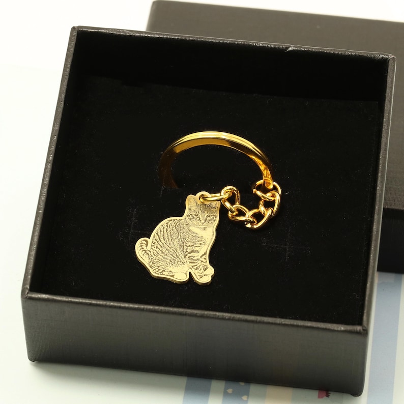 May include: Gold-colored keychain with a small, engraved image of a cat. The keychain is in a black gift box.