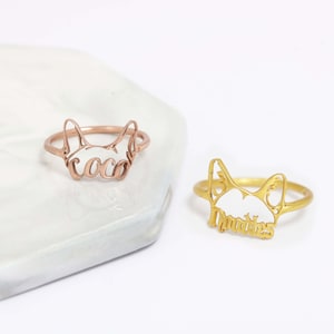 K&ouml;nnte beinhalten: Zwei Metallringe mit einem ausgeschnittenen Katzenkopf-Design. Ein Ring ist ros&eacute;goldfarben mit dem Namen coco in Schreibschrift, der andere ist goldfarben mit dem Namen Mookie in Schreibschrift, der in die Katzenohren integriert ist.