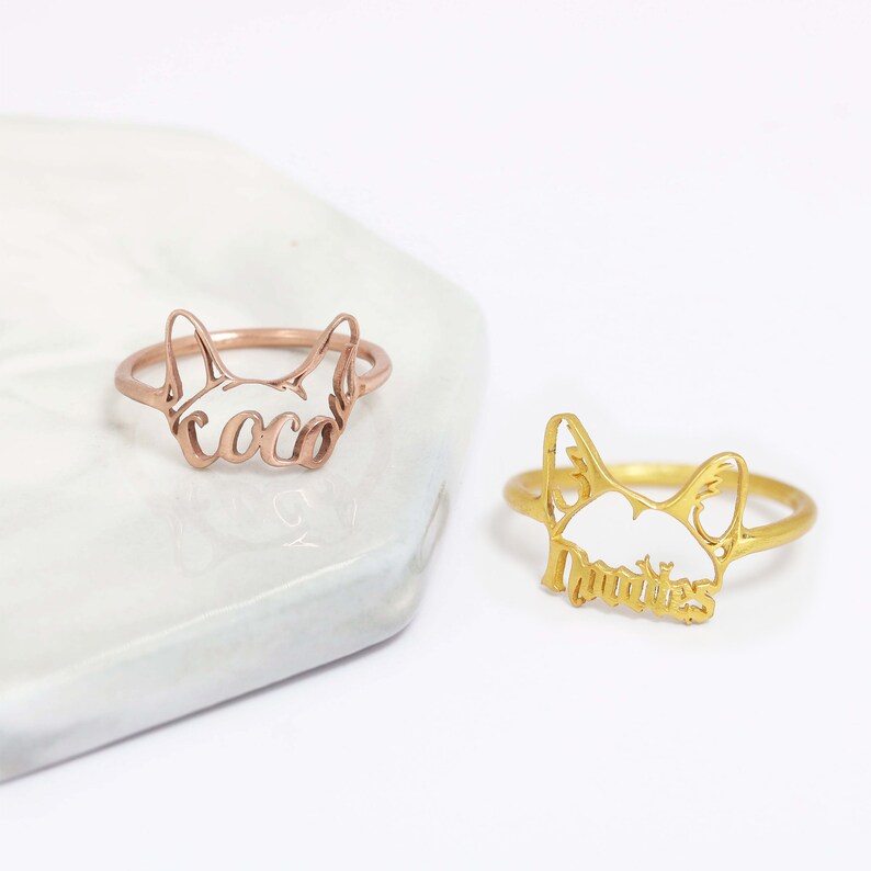 K&ouml;nnte beinhalten: Zwei Metallringe mit einem ausgeschnittenen Katzenkopf-Design. Ein Ring ist ros&eacute;goldfarben mit dem Namen coco in Schreibschrift, der andere ist goldfarben mit dem Namen Mookie in Schreibschrift, der in die Katzenohren integriert ist.