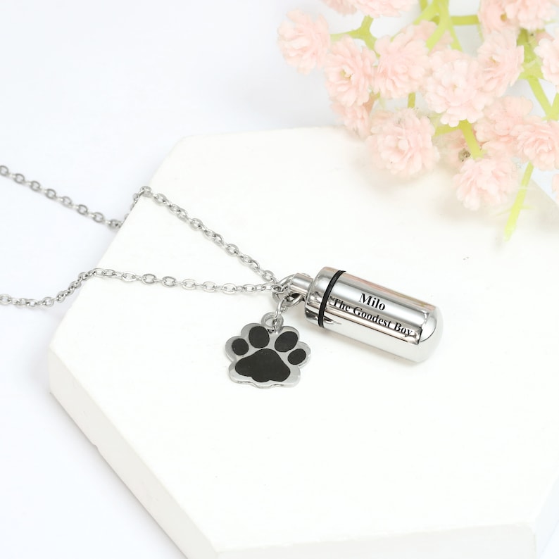 Op de afbeelding: Een zilverkleurige metalen urn-hanger ketting met een cilindrische kamer gegraveerd met de tekst Milo The Goodest Boy en een kleine bungelende bedel in de vorm van een pootafdruk met een zwart midden, aan een fijne ketting.