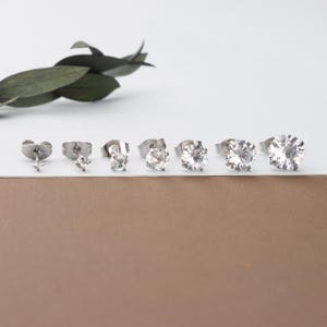 May include: A collection of six pairs of silver-toned stud earrings. The earrings vary in size, with the smallest featuring a star-shaped design and the others showcasing round, clear gemstones. The earrings are displayed on a white surface.