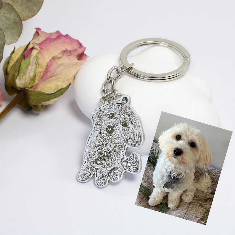 May include: Silver keychain with a photo of a white dog engraved on it. The keychain is attached to a silver key ring.