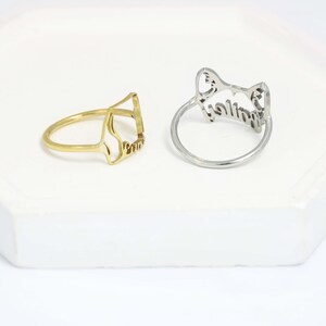 K&ouml;nnte beinhalten: Zwei Ringe aus Metall, einer goldfarben und einer silberfarben. Jeder Ring verf&uuml;gt &uuml;ber ein ausgeschnittenes Design in Form von Katzenohren an der Oberseite, wobei ein Name in Schreibschrift in die Ringstruktur integriert ist. Die Ringe sind d&uuml;nn.