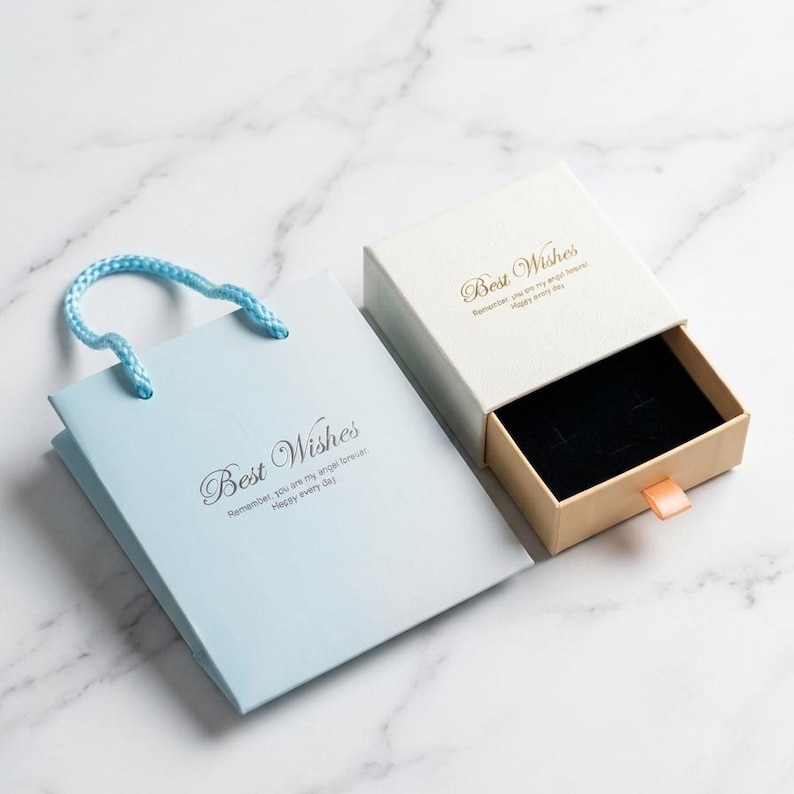 Op de afbeelding: Een lichtblauwe papieren geschenktas met een blauw koordhengsel en een bijpassend wit schuifdoosje voor sieraden. Beide items zijn voorzien van de tekst Best Wishes en de zin Remember, you are my angel forever. Happy every day in goud en zwart.