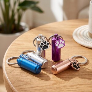 Op de afbeelding: Vier cilindervormige metalen urn-sleutelhangers in blauw, zilver, paars en ros&eacute;goud. Elke sleutelhanger is voorzien van een kleine bedel in de vorm van een pootafdruk met een contrasterend midden. De houders hebben een schroefdeksel en een ring.