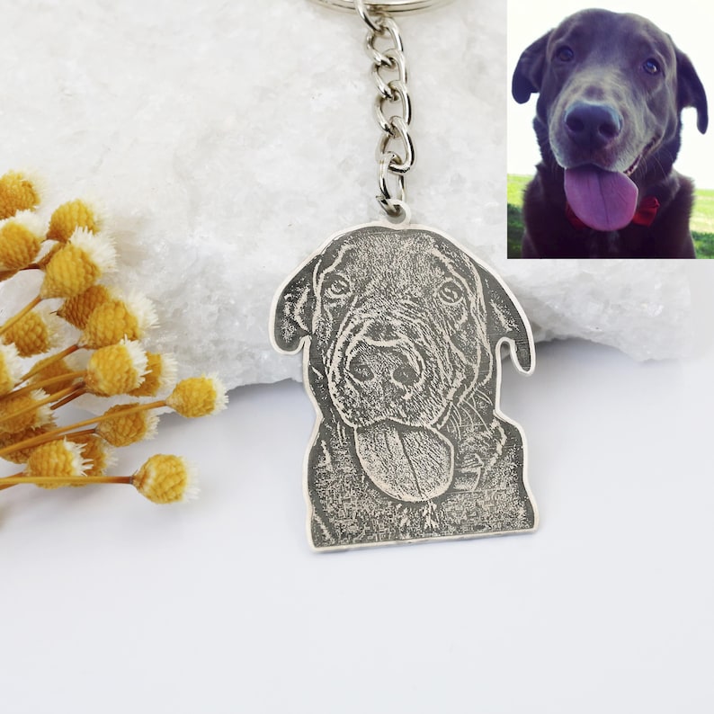 May include: Silver keychain with a black and white engraved image of a dog's head. The dog is sticking its tongue out and has a happy expression.