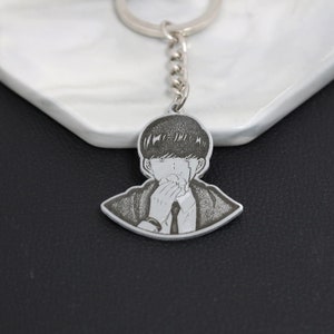May include: A silver keychain featuring a black and white engraved image of a person with short hair, wearing a suit and tie, covering their mouth with their hand.