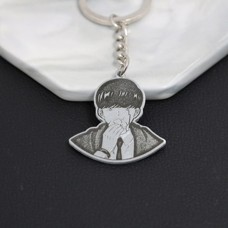 May include: A silver keychain featuring a black and white engraved image of a person with short hair, wearing a suit and tie, covering their mouth with their hand.