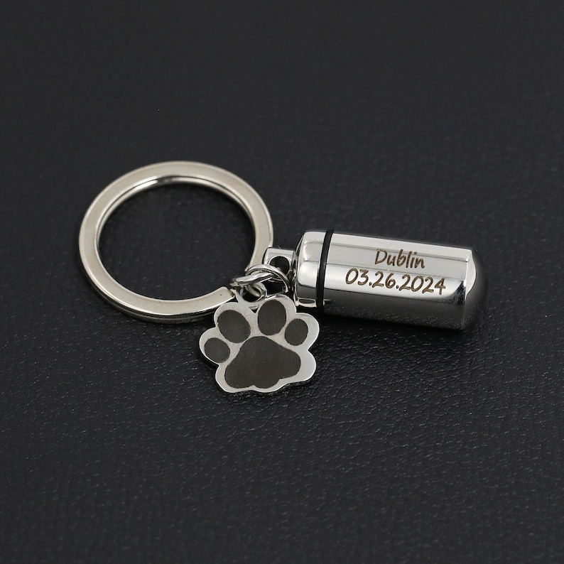 Op de afbeelding: Een zilverkleurige metalen sleutelhanger met een cilindervormige capsulehanger gegraveerd met de tekst Dublin en 03.26.2024, vergezeld van een kleine bedel in de vorm van een pootafdruk met zwarte vulling, bevestigd aan een ronde sleutelring.