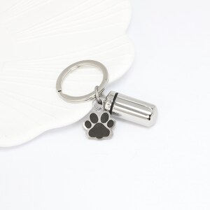 Op de afbeelding: Een zilverkleurige metalen sleutelhanger met een cilindervormige capsulehanger met een zwarte rubberen afdichting en een kleine, platte bedel in de vorm van een pootafdruk met een zwart midden. Inclusief een ronde ring voor sleutels.