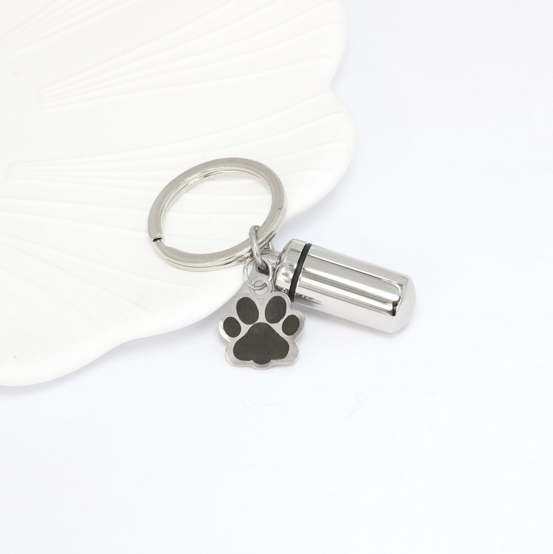 Op de afbeelding: Een zilverkleurige metalen sleutelhanger met een cilindervormige capsulehanger met een zwarte rubberen afdichting en een kleine, platte bedel in de vorm van een pootafdruk met een zwart midden. Inclusief een ronde ring voor sleutels.