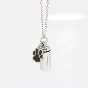 Op de afbeelding: Een zilverkleurige cilindervormige hanger aan een ketting met een kleine, bungelende bedel in de vorm van een pootafdruk. De pootafdrukbedel heeft een zwart midden en de hanger hangt aan een fijne zilverkleurige ketting met ronde schakels.