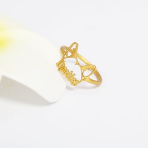 K&ouml;nnte beinhalten: Ein goldfarbener Ring mit einem ausgeschnittenen Design in Form einer Katzenkopf-Silhouette. Die Katzenohren sind spitz und der untere Teil der Ringvorderseite enth&auml;lt das Wort "Amities" in einer kursiven Schriftart. Die Ringschiene ist d&uuml;nn.