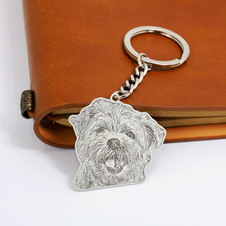 May include: Silver keychain with a photo engraved of a dog's face. The dog has a white and black coat and is smiling.