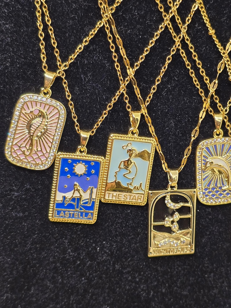 May include: Five gold necklaces with different pendants featuring colorful enamel and rhinestones. The pendants are rectangular and feature images of a seahorse, a woman with a star, a woman with a moon, a woman with a sun, and a dolphin. The pendants have the words "LASTELLA", "THE STAR", and "KNIGHT OF SWORDS" written on them.