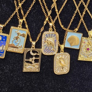 May include: A set of gold chain necklaces with various pendants featuring tarot card designs. The pendants are rectangular and have a blue and white enamel finish with gold accents. The pendants feature images of a star, a knight of cups, a seashell, a bird, and a wheel of fortune.