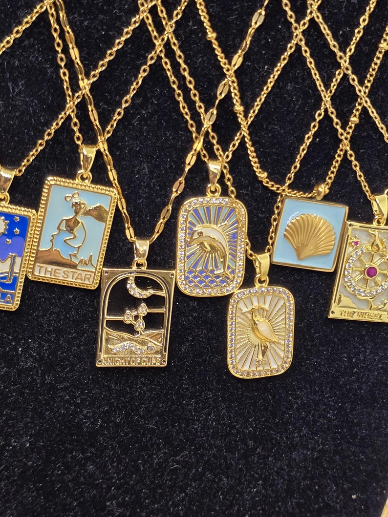 May include: A set of gold chain necklaces with various pendants featuring tarot card designs. The pendants are rectangular and have a blue and white enamel finish with gold accents. The pendants feature images of a star, a knight of cups, a seashell, a bird, and a wheel of fortune.