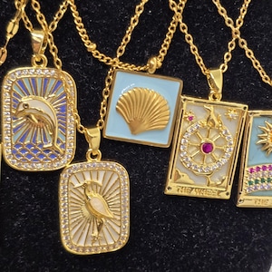 May include: A set of gold necklaces with various pendants featuring a dolphin, a seashell, a wheel with a red jewel, and a bird. The pendants are surrounded by small, clear stones and have a blue and white enamel background.