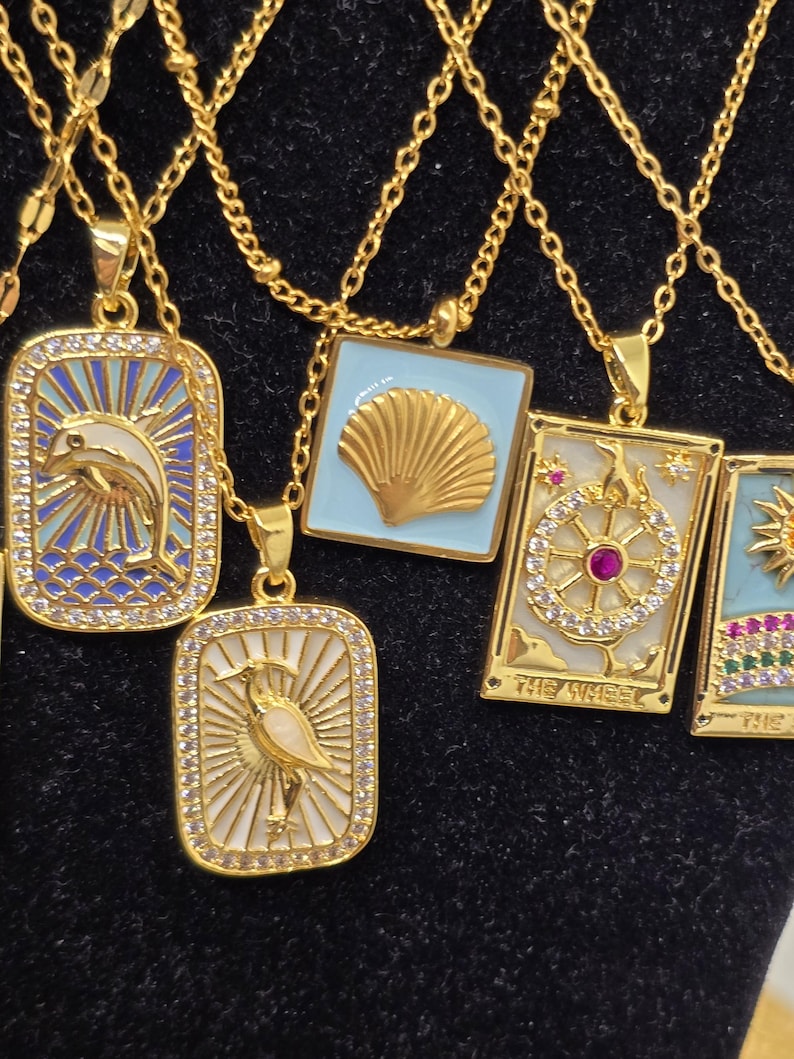 May include: A set of gold necklaces with various pendants featuring a dolphin, a seashell, a wheel with a red jewel, and a bird. The pendants are surrounded by small, clear stones and have a blue and white enamel background.