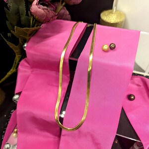 Könnte beinhalten: Eine goldene Kettenkette auf einem rosa Satinband. Die Kette ist ein schlichtes, flaches Design.