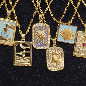 May include: A set of nine gold necklaces with different pendants. Each pendant is a square with a different design, including a seashell, a dolphin, a bird, and a lighthouse. The pendants are all surrounded by small, clear rhinestones.