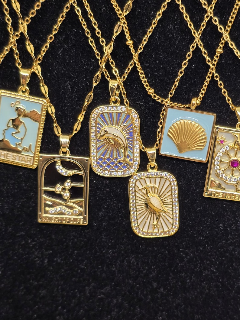 May include: A set of nine gold necklaces with different pendants. Each pendant is a square with a different design, including a seashell, a dolphin, a bird, and a lighthouse. The pendants are all surrounded by small, clear rhinestones.