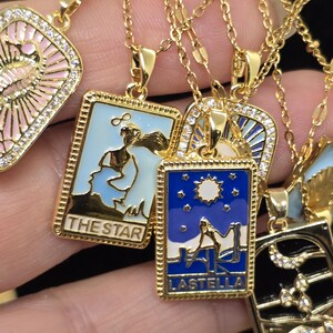 May include: Gold necklaces with pendants featuring colorful enamel designs and rhinestones. The pendants are shaped like rectangular cards with text that reads "The Star" and "Lastella".
