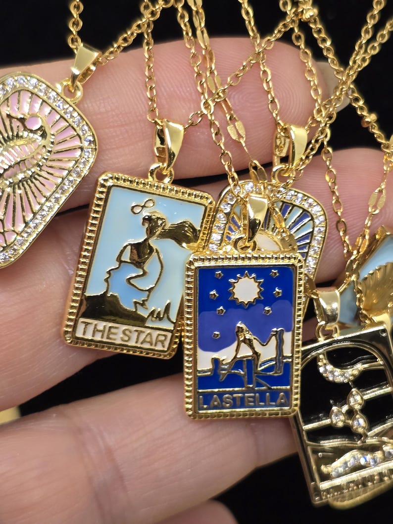 May include: Gold necklaces with pendants featuring colorful enamel designs and rhinestones. The pendants are shaped like rectangular cards with text that reads "The Star" and "Lastella".