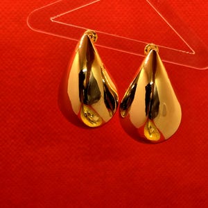 Könnte beinhalten: Ein Paar goldene Ohrringe in Tropfenform. Die Ohrringe hängen an einem durchsichtigen Plastikaufhänger vor einem roten Hintergrund.