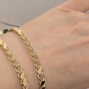 Könnte beinhalten: Zwei goldene Kettenarmbänder mit einem blattförmigen Gliederdesign. Die Armbänder werden am Handgelenk einer Person getragen.