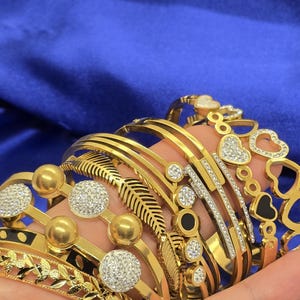 Könnte beinhalten: Ein Set goldfarbener Armbänder mit verschiedenen Designs, darunter ein Blatt, ein Herz und eine Reihe kleiner Kreise. Einige der Armbänder sind mit klaren Strasssteinen verziert.