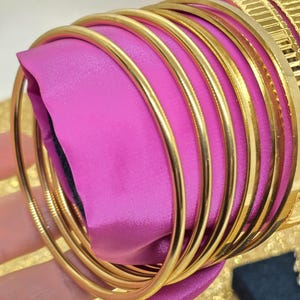 Könnte beinhalten: Ein Satz goldener Armreifen, gestapelt und auf einem rosa Stoffhintergrund angeordnet. Die Armreifen sind glatt und glänzend, mit einer polierten Oberfläche.