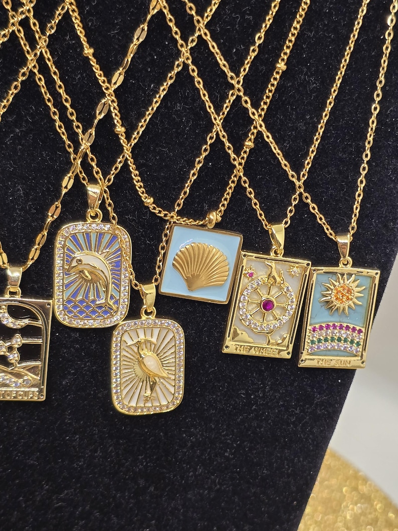 May include: A set of gold necklaces with various pendants featuring colorful enamel and rhinestones. The pendants include a dolphin, a seashell, a tarot card with the Wheel of Fortune, and a tarot card with the Sun.