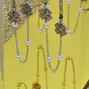 Könnte beinhalten: Silberfarbene Ohrringe mit einem Kleeblatt-Design. Jeder Ohrring hat einen Kleeblatt-Anhänger, eine Kette und kleine weiße Perlen. Die Ohrringe werden vor einem goldenen Hintergrund mit Glitzer präsentiert.