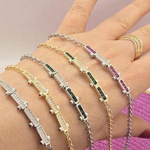 Könnte beinhalten: Eine Sammlung zarter Armbänder in Gold- und Silbertönen, jedes mit einem rechteckigen Mittelstück, das mit funkelnden Edelsteinen besetzt ist. Die Edelsteine umfassen klare, grüne und rote Farbtöne und verleihen dem Schmuck einen Hauch von Farbe.