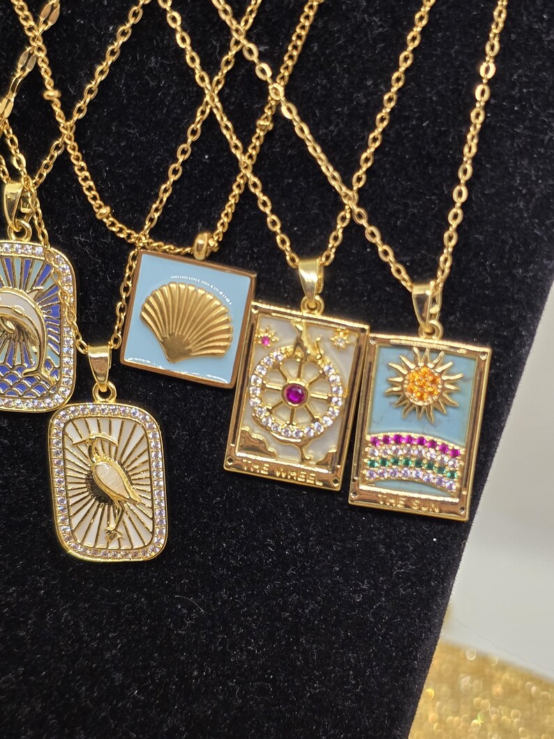 May include: Four gold necklaces with different pendants. The pendants are square and feature a seashell, a sun, a wheel, and a bird. Each pendant is decorated with rhinestones and colorful gems.